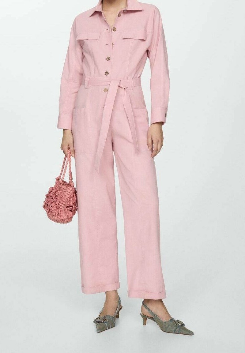 Femme portant une combinaison rose ceinturée avec poches avant et boutons, tenant un sac à main rose texturé et portant des talons pointus gris.