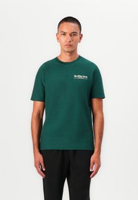 Mørkegrøn kortærmet t-shirt lavet af glat bomuld, med et lille hvidt grafisk logo på venstre bryst. Lige kant, afslappet pasform.