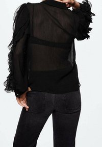 Blusa nera trasparente con maniche a volant, caratterizzata da un colletto e un retro traslucido. Abbinata a jeans scuri e aderenti.