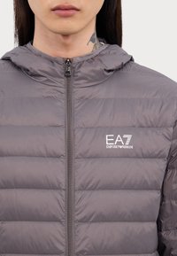 Grå pufferjakke med hette, med glidelås og vannrett quiltet design. EA7 Emporio Armani-logo på brystet. Glatt tekstur.
