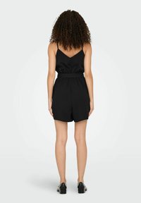 Combinaison noire avec de fines bretelles spaghetti, un dos en V, une taille élastique et des shorts amples. Le tissu semble lisse avec une finition mate.