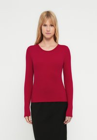 Top rosso a maniche lunghe con trama a coste e scollo rotondo. Taglio aderente con leggero effetto elasticizzato. Abbinato a una gonna nera. Design minimalista.