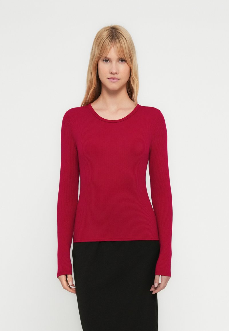 Top rosso a maniche lunghe con trama a coste e scollo rotondo. Taglio aderente con leggero effetto elasticizzato. Abbinato a una gonna nera. Design minimalista.