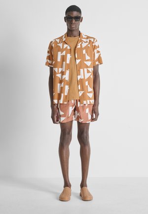Man die een bruin patroon kort mouwen shirt draagt, bijpassende shorts, beige T-shirt, beige schoenen en zwarte zonnebril, staand tegen een witte achtergrond.