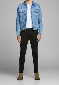 Chaqueta de mezclilla azul claro con dos bolsillos en el pecho, combinada con pantalones negros ajustados y botas de tobillo beige, contra un fondo neutral.