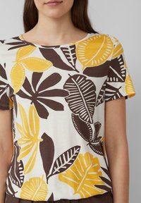 T-shirt à manches courtes en tissu crème, avec un grand imprimé botanique comprenant des feuilles jaunes et brunes, un col arrondi et une coupe décontractée.