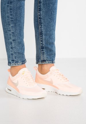 Person trägt helle rosa Nike-Sneaker und blaue Jeans, steht auf einem weißen Boden vor einem schlichten Hintergrund.