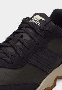 Μαύρο και ελαιδί αθλητικό παπούτσι Sorel με χοντρές κορδόνια, ραμμένες λεπτομέρειες και λευκή ελαστική σόλα με καφέ τόνους.