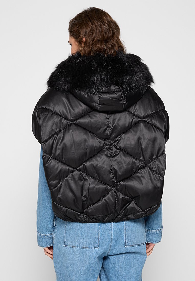 Donna che indossa un gilet imbottito nero con cappuccio foderato in pelliccia sopra una giacca di jeans blu, vista da dietro.