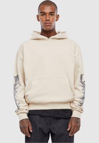 Beige hættetrøje med kængurulomme, prydet med detaljerede grafiske designs i sølv på ærmerne. Blødt stof, afslappet pasform.