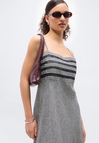 Νεαρή γυναίκα που φοράει ασπρόμαυρο φόρεμα με καρό και δαντέλα, σκούρα γυαλιά ηλίου, ασημένια σκουλαρίκια και βραχιόλι, κρατώντας μια μπορντό τσάντα ώμου.