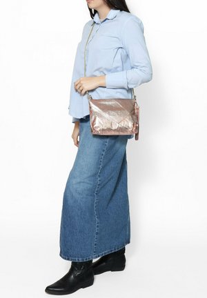 Borsa metallizzata color rosé oro con un design strutturato, dettagli trapuntati e una tracolla a catena. Indossata in stile crossbody contro una camicia azzurra.