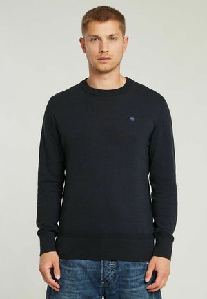 Maglione lavorato a maglia blu navy con scollo rotondo, maniche lunghe e polsini a costine, con un piccolo logo sul petto. Indossato con jeans in denim.