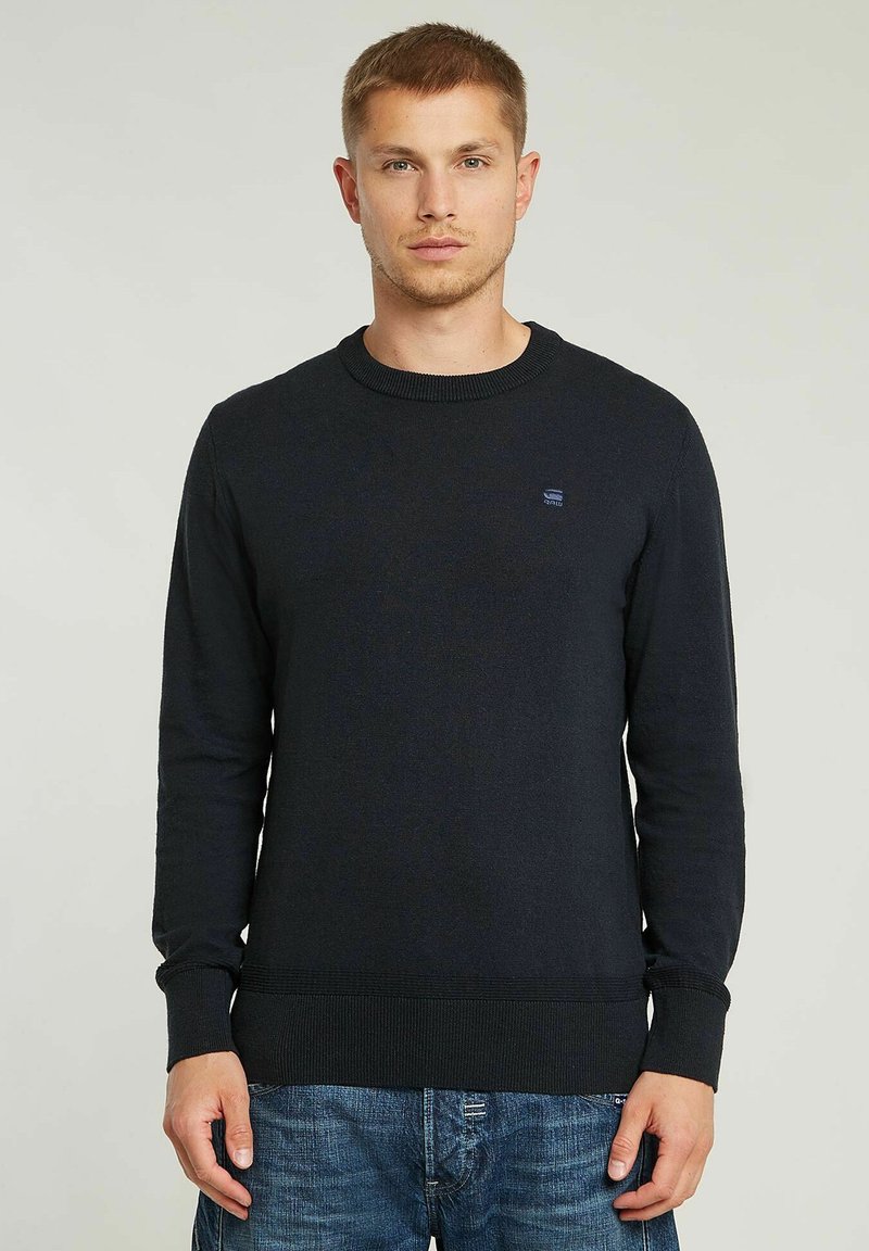 Maglione lavorato a maglia blu navy con scollo rotondo, maniche lunghe e polsini a costine, con un piccolo logo sul petto. Indossato con jeans in denim.