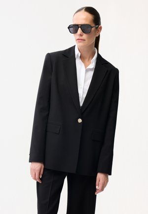 Vrouw die een oversized zwart blazer met gouden knoop, wit overhemd, zwarte broek en grote zwarte zonnebril draagt, staand tegen een witte achtergrond.