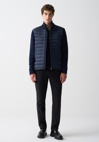 Mladý muž v tmavomodrom prešívanom veste cez tmavú košeľu s dlhým rukávom, čiernych nohaviciach a čiernych mokasínach, stojí pred jednoduchým pozadím.