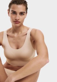 Jonge vrouw met een lichte huid en bruin haar dat naar achteren is gekamd, draagt een naadloos, mouwloos crop top in nude kleur tegen een effen witte achtergrond.