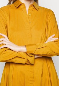 Koszula z długim rękawem i kołnierzykiem w musztardowym kolorze, wykonana z gładkiego materiału. Posiada guziki z przodu i luźny krój.