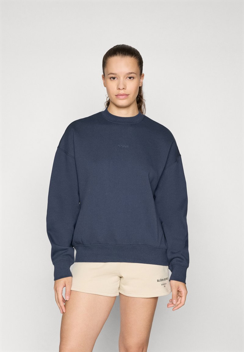 Navyblaues Sweatshirt aus weichem Material, mit Rundhalsausschnitt, überschnittenen Schultern und kleinem Logo auf der Brust. Kombiniert mit beigen Shorts.