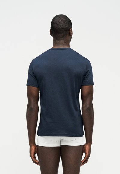 Άνδρας με την πλάτη, φοράει απλό σκούρο μπλε κοντομάνικο και λευκά εσώρουχα, σε απλό ανοιχτόχρωμο φόντο.