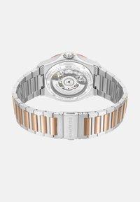 Srebrny i różowozłoty zegarek ze stali nierdzewnej z skomplikowanym mechanicznym tyłem, z teksturowanym paskiem i okrągłą kopertą.