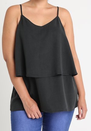 Frau trägt ein locker sitzendes, schwarzes, doppellagiges ärmelloses Oberteil mit dünnen Trägern und blaue Jeans, steht vor einem schlichten Hintergrund.