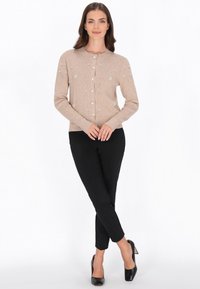 Beige kofta med knäppning framtill, smyckad med små blommönster och paljetter, i kombination med svarta, figurnära byxor och svarta klackar.