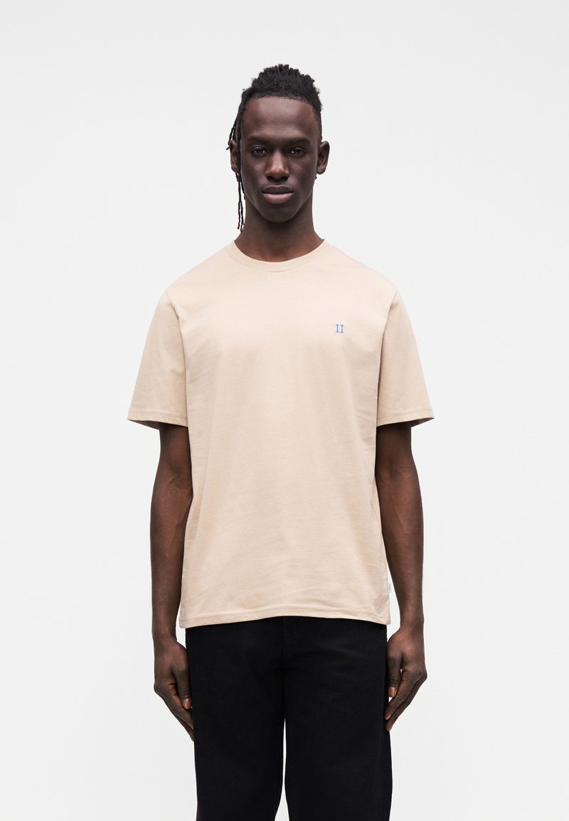 Jasnobeżowy bawełniany t-shirt z krótkimi rękawami i okrągłym dekoltem. Na piersi małe niebieskie logo "II". Noszony z czarnymi spodniami.