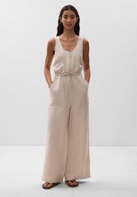 Beige Leinen-Jumpsuit mit ärmellosem Design, weiten Beinen, Kordelzug in der Taille und seitlichen Taschen. Modell trägt flache Sandalen.