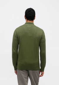 Πουλόβερ από πλεκτό ελιάς με ριμπ γιακά, μακριά μανίκια και άνετη εφαρμογή, συνδυασμένο με ανοιχτό γκρι παντελόνι.