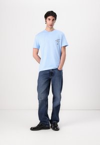 T-shirt en coton bleu clair à manches courtes, avec un design de poche poitrine. Associé à un jean bleu foncé à coupe décontractée et des chaussures noires.