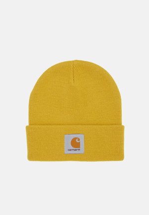 Gelbe Strickmütze mit umgeschlagenem Bündchen und quadratischem weißen Aufnäher, der das orangefarbene Carhartt-Logo mittig auf dem Bündchen zeigt.