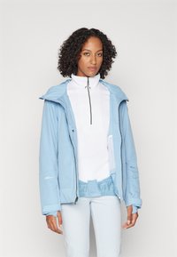 Hellblauer Zip-Hoodie mit glatter Textur und Seitentaschen, getragen über einem weißen hochkragen Oberteil und hellen Hosen.