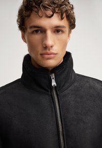 Giacca nera in pelle sintetica con colletto alto, dotata di chiusura con zip metallica e finitura testurizzata. Design semplice e moderno, privo di fantasie.