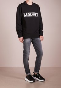 Sudadera negra con texto blanco "JUST", cuello redondo y puños de canalé, combinada con jeans grises descoloridos y zapatillas deportivas negras.