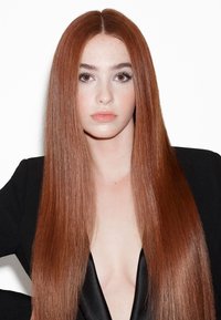 Jeune femme aux longs cheveux raides rouges et aux yeux marron portant un blazer noir sur un haut plongeant, posant devant un fond blanc.