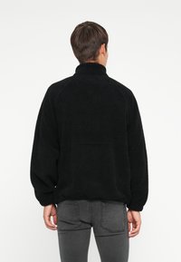 Svart fleecepullover med høy krage, raglanermer, og strukturell finish. Mykt materiale og avslappet passform, kombinert med mørkegrå jeans.