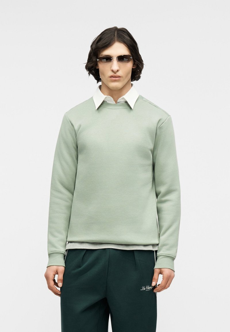 Νέος άνδρας με ανοιχτοπράσινο φούτερ, λευκό πουκάμισο με γιακά από κάτω, σκούρα πράσινα παντελόνια και γυαλιά ηλίου, στέκεται μπροστά σε απλό φόντο.