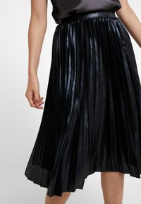Gonna plissettata nera realizzata in materiale lucido, presenta una vita ampia e scende sotto il ginocchio, mettendo in mostra una texture liscia e linee pulite.