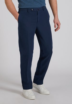 Mężczyzna ubrany w granatowe spodnie o prostych nogawkach, białe trampki i niebieską koszulę, z jedną ręką w kieszeni na jednolitym tle.