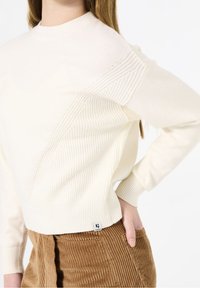 Krämfärgad stickad tröja med ett strukturerat diagonalmönster och ribbade ärmmuddar, kombinerad med bruna kordflorsbyxor. Litet varumärkesmärke synligt.