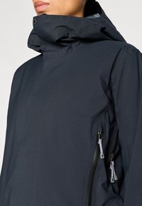 Jachetă impermeabilă de culoare bleumarin închis, cu guler înalt, fermoare pe părțile laterale și textură fină. Dispune de o glugă reglabilă și un design minimalist.