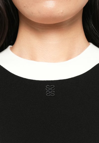 Close-up van de nek en onderkant van het gezicht van een persoon die een zwart sweatshirt met een witte kraag en een klein geborduurd zwart logo draagt.