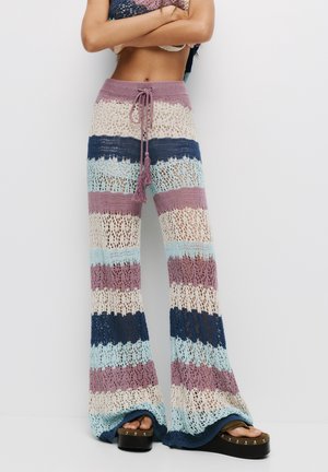 Osoba ubrana w wielokolorowe, paskowane spodnie szydełkowe z wiązanym pasem i sandały na platformie, stojąca na tle jednolitego białego tła.