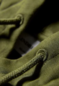 Olivgrön tyg med en slät textur, som visar upp flätad dragsko och hållets detaljer. Etikett delvis synlig.