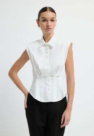 Blouse blanche sans manches avec un col pointu, fermeture à boutons sur le devant et coupe structurée, associée à un pantalon noir taille haute.