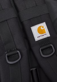 Sort nylon rygsæk med justerbare stropper, rektangulær form og et hvidt og orange Carhartt-logo patch. Holdbarhed er fremhævet i designet.