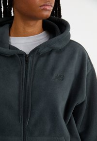 Tmavě šedá fleecová mikina s kapucí a zipem, se strukturovaným povrchem, se černým vyšitým logem a podšívkou kapuce. Nositá přes světle šedý top.