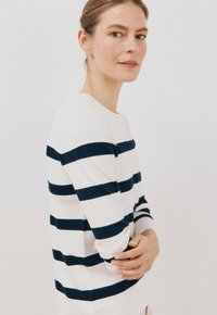 Maglione bianco con righe orizzontali blu navy, scollo rotondo, maniche lunghe, texture liscia e vestibilità aderente, indossato da un modello.