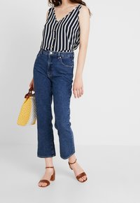 Jeans bleus taille haute, longueur raccourcie. Haut sans manches rayé bleu marine et blanc avec boutons. Sandales à talons blocs marron avec lanières. Sac fourre-tout jaune.